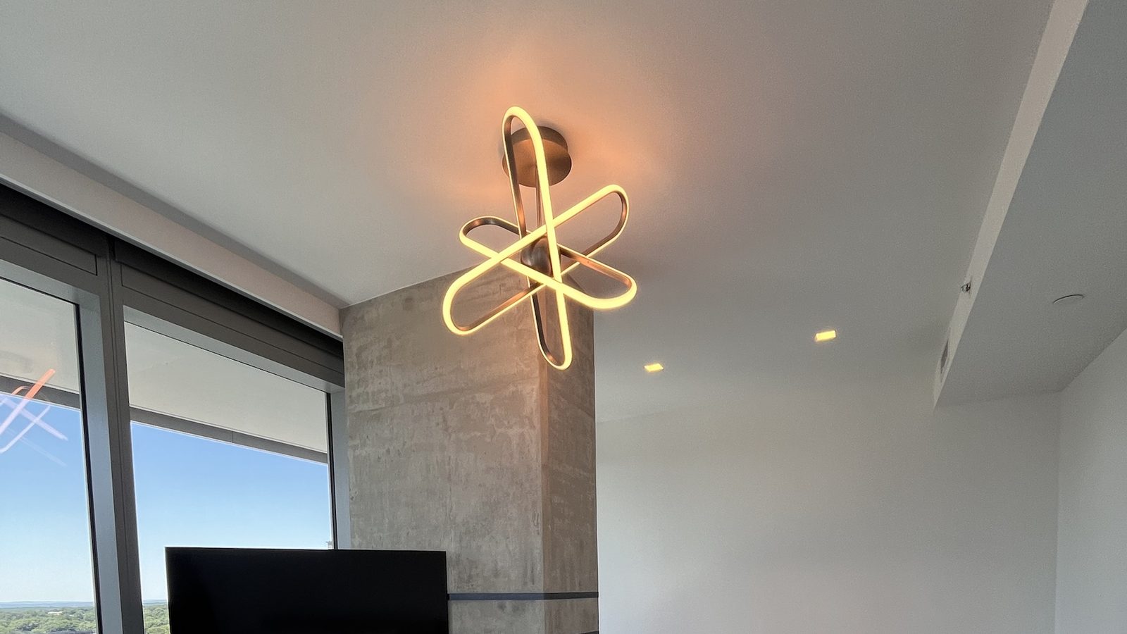 Modern LED ring ceiling fixture installed above a concrete column with a wall-mounted TV in a high-rise bedroom — Argos AV light fixture install